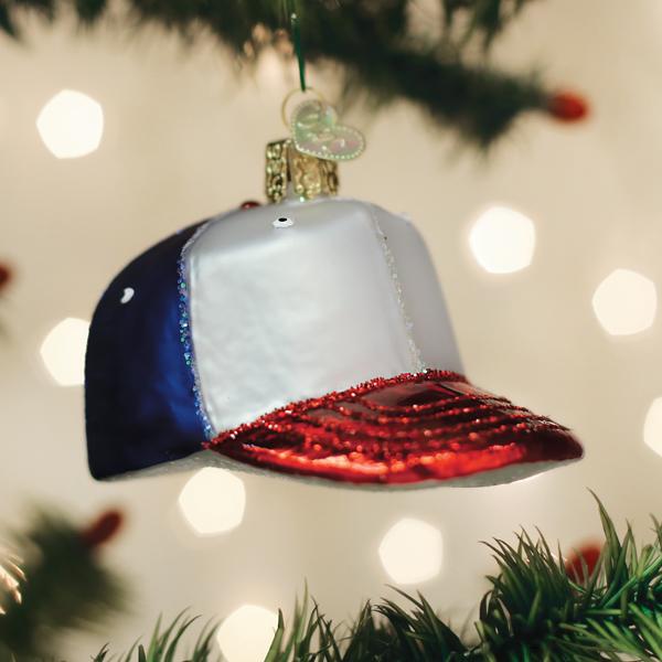 A Christmas tree ornament shaped like a red, white, and blue baseball cap hangs among green pine branches with blurred lights in the background.