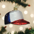 A Christmas tree ornament shaped like a red, white, and blue baseball cap hangs among green pine branches with blurred lights in the background.