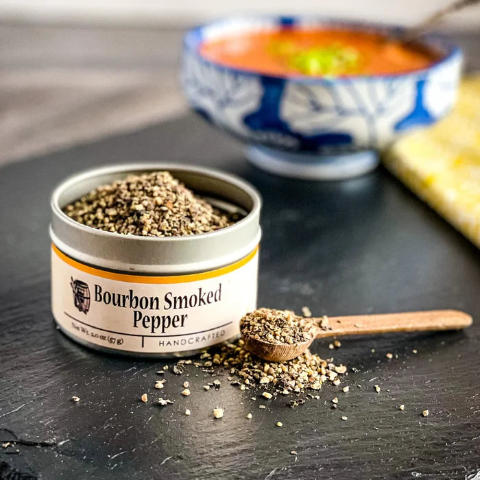 Bourbon Smoked Pepper container with a wooden spoon on a dark surface, with a blurred bowl of soup in the background.