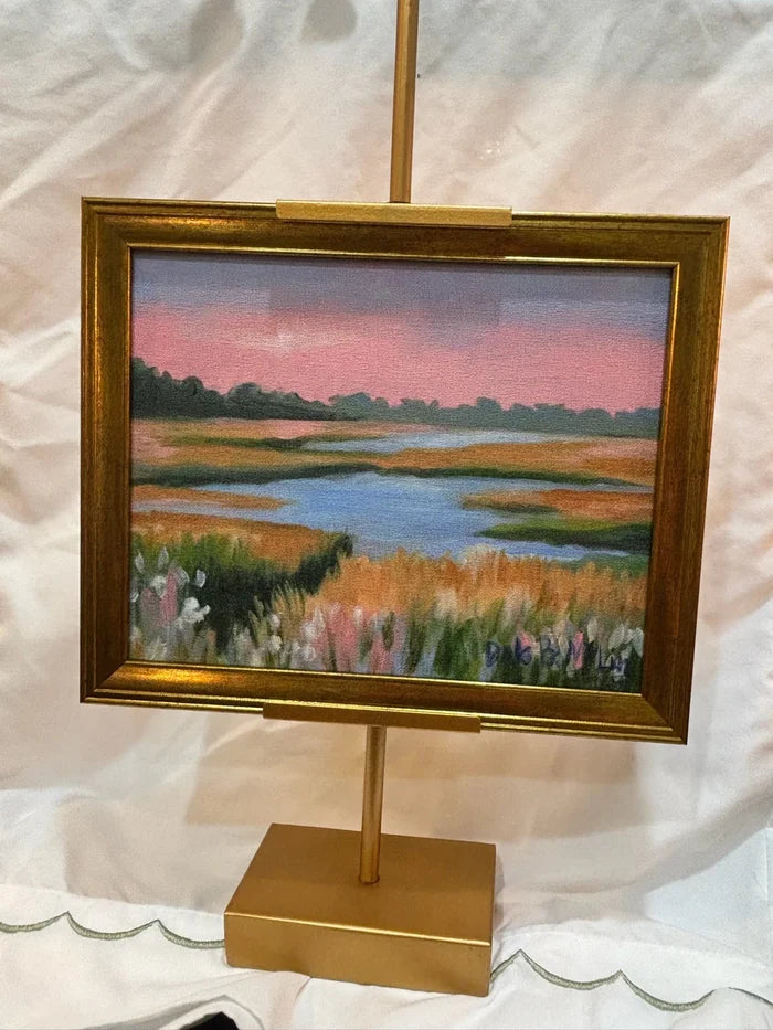 Painting of a landscape with a lake and fields on a gold easel against a white background