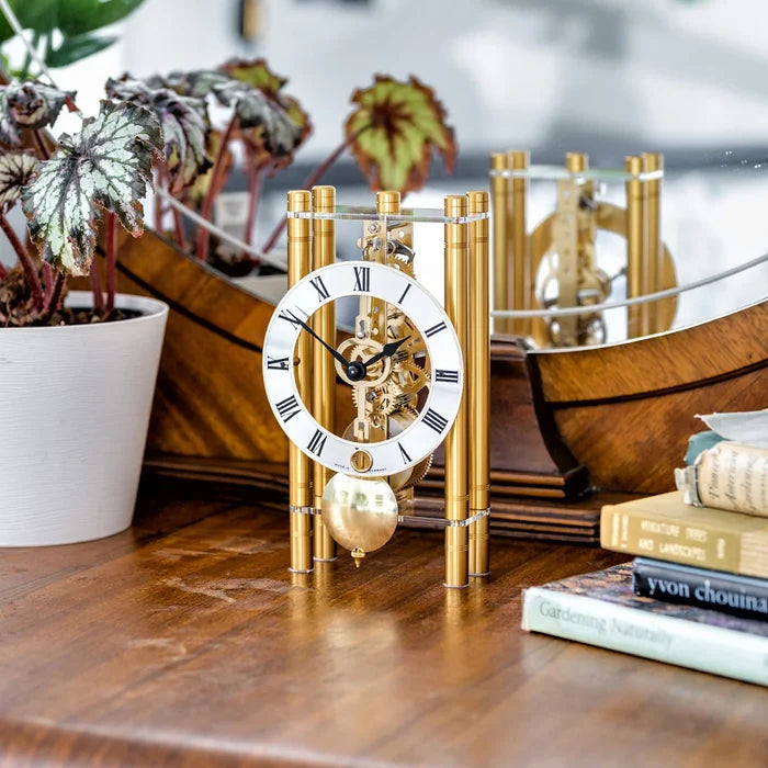 Gold mechanical clock on a wooden surface with books and plants in the background