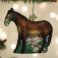 A brown horse-shaped Christmas ornament with a blue bridle hangs on a tree branch, surrounded by green glittery grass and blurred white lights in the background.