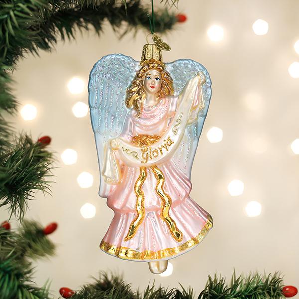 A glass Christmas tree ornament shaped like an angel with blue wings and a pink and gold dress, holding a banner that reads Gloria, hanging from a branch with blurred lights in the background.