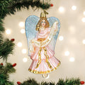 A glass Christmas tree ornament shaped like an angel with blue wings and a pink and gold dress, holding a banner that reads Gloria, hanging from a branch with blurred lights in the background.