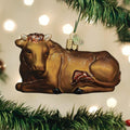 A brown glass cow ornament with glitter details hangs from a green Christmas tree branch, with soft white holiday lights glowing in the blurred background.