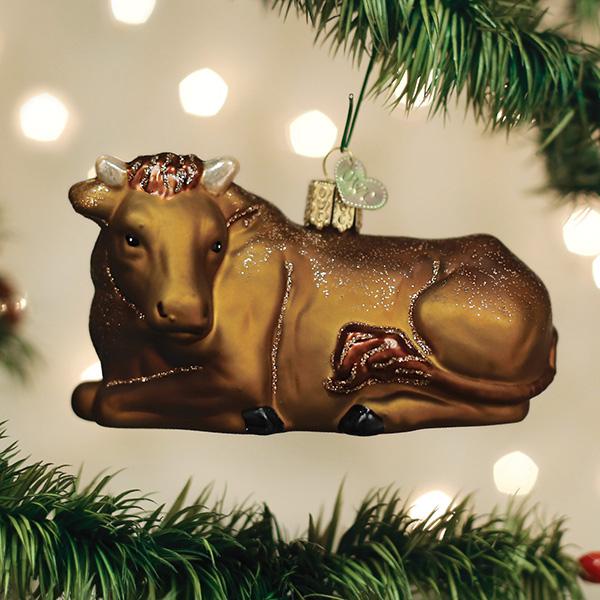 A brown glass cow ornament with glitter details hangs from a green Christmas tree branch, with soft white holiday lights glowing in the blurred background.