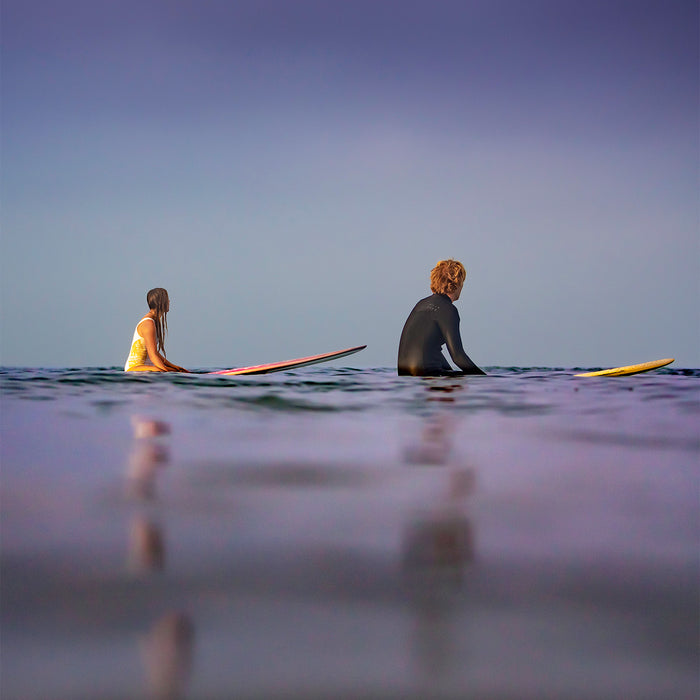 Two surfers seated on surfboards in calm purple-toned water at dusk, shot at sea level in the fine art photograph PATIENT COUPLE by Craig Hubbard.