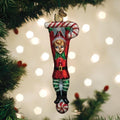 A festive Christmas tree ornament shaped like a cheerful elf in a red outfit, hanging upside down from a glittery candy cane, with twinkling lights and green tree branches in the background.