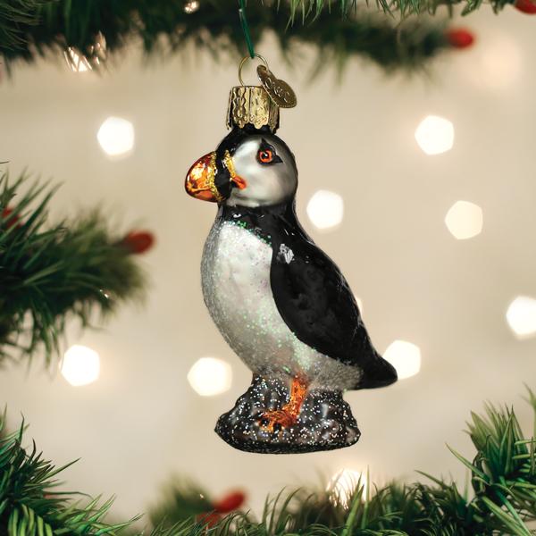 A glass puffin ornament hangs from a Christmas tree branch with blurred lights in the background.