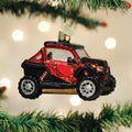 A red and black off-road vehicle ornament hangs on a Christmas tree, with blurred white lights in the background.