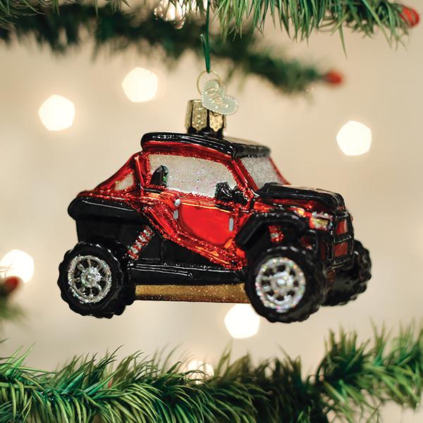 A red and black off-road vehicle ornament hangs on a Christmas tree, with blurred white lights in the background.