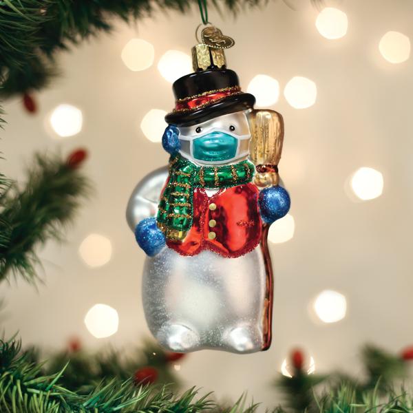 A glass snowman ornament wearing a red vest, green scarf, black hat, and a blue face mask hangs on a Christmas tree, with twinkling lights blurred in the background.