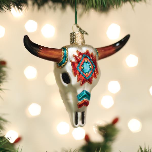 A Christmas tree ornament shaped like a decorated bull skull with colorful patterns hangs from a branch, with soft, out-of-focus holiday lights in the background.