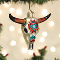 A Christmas tree ornament shaped like a decorated bull skull with colorful patterns hangs from a branch, with soft, out-of-focus holiday lights in the background.