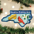 A Christmas tree ornament shaped like North Carolina features the words â€œNorth Carolina,â€_x009d_ a red cardinal, a white dogwood flower, and a yellow airplane, hanging among green pine branches with white lights.