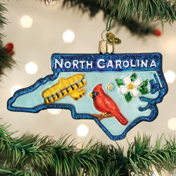 A Christmas tree ornament shaped like North Carolina features the words â€œNorth Carolina,â€_x009d_ a red cardinal, a white dogwood flower, and a yellow airplane, hanging among green pine branches with white lights.