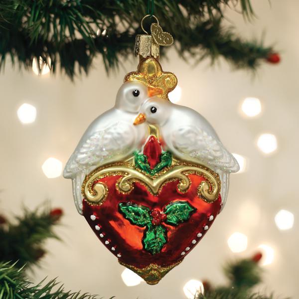 A Christmas tree ornament shaped like two white doves perched on a red heart decorated with holly leaves and berries, with gold accents and a gold hanging hook. Out-of-focus lights shine softly in the background.
