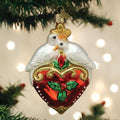 A Christmas tree ornament shaped like two white doves perched on a red heart decorated with holly leaves and berries, with gold accents and a gold hanging hook. Out-of-focus lights shine softly in the background.