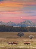 Fine art photograph of horses grazing in a open field at dusk, with a glowing moon above pastel pink and orange skies and snow-capped mountains in the distance.