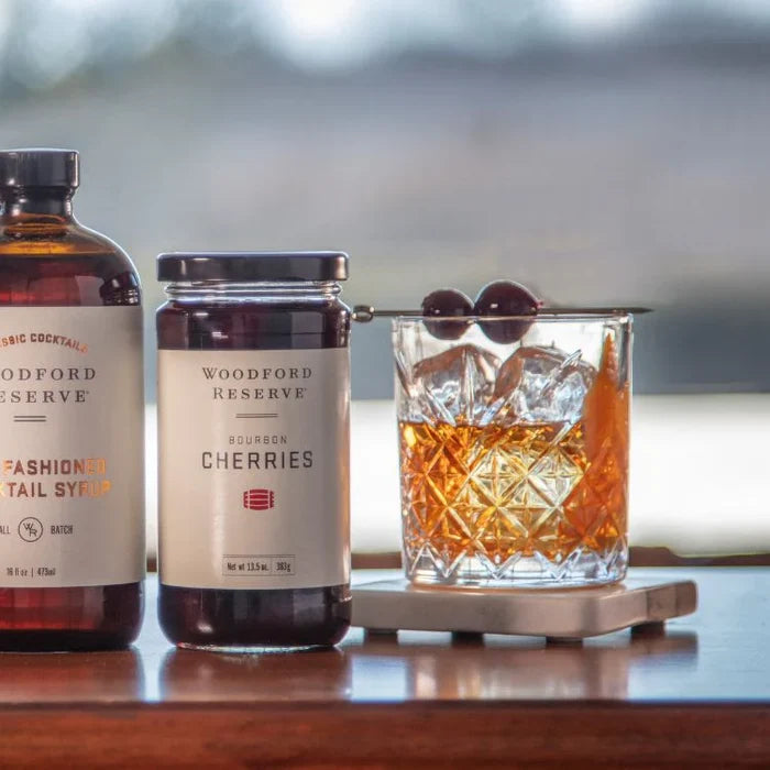 Woodford Reserve cocktail ingredients including a bottle, jar, and glass with a drink on a wooden surface.